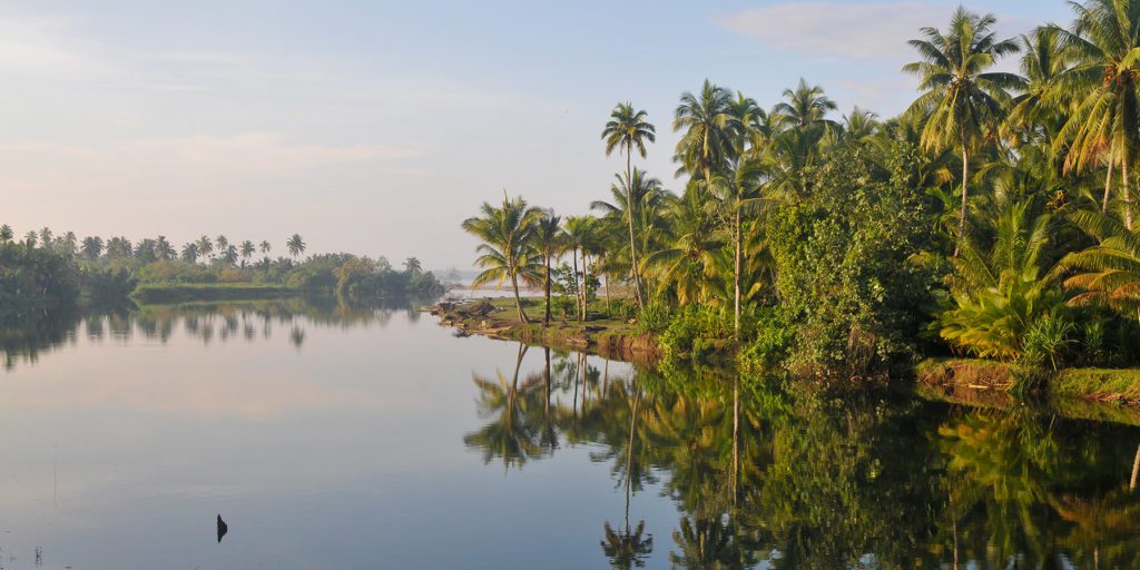 Tranquil river reflecting cloudless sky, framed by tropical vegetation on the side. Sri Lanka
