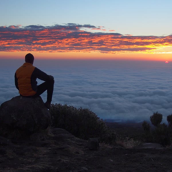 Trekker sitting on a rock, overlooking the sun rise over the top of the clouds on Mount Kilimanjaro