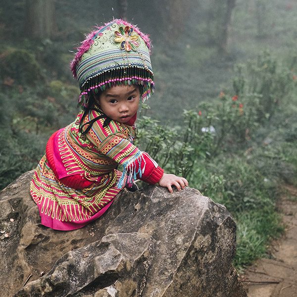 Little Hmong child in bright traditional dress sitting on a rock. Vietnam