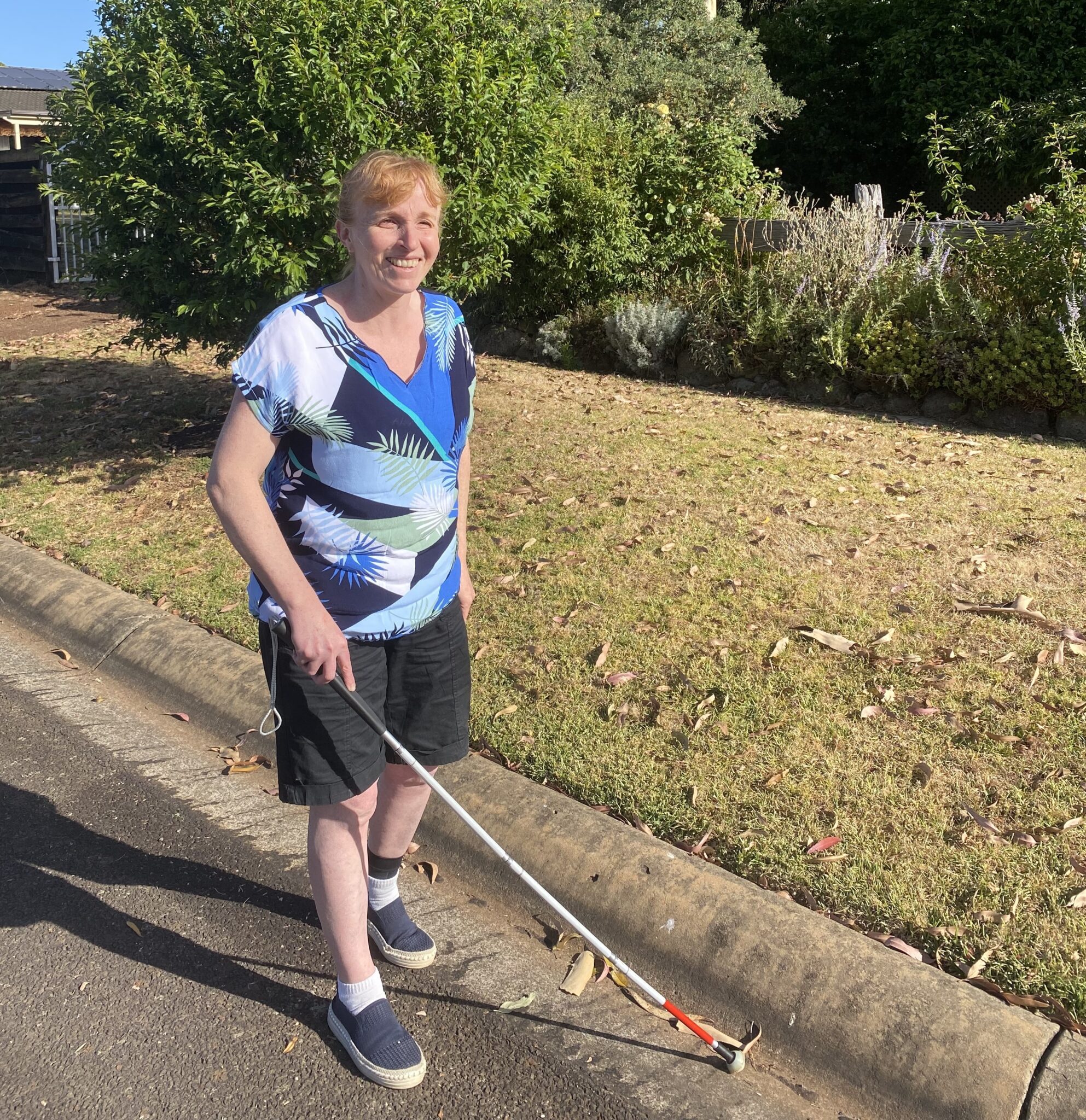 Image of Vision Australia client Beth smiling whilst walking down the road with her white cane.