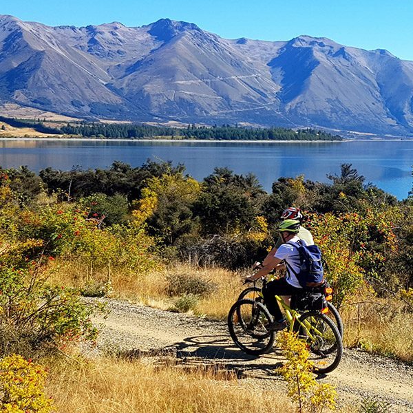 Cyclists side by side along a rocky path with the lake and mountain rising up in the background